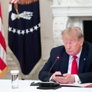Donald Trump looks at a smartphone while sitting at a desk.